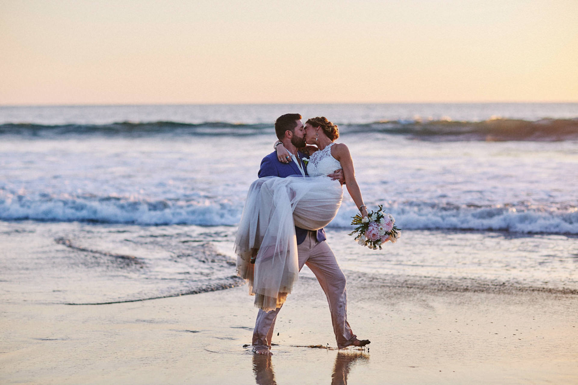 Bride and groom kissing at sunset during a Costa Rica elopement on the beach, groom carrying bride by the ocean after an intimate destination wedding